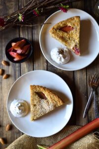 An overview of two plates of sliced Bakewell Tart | Roasted Rhubarb with fresh whipped cream, almonds, a small bowl of roasted rhubarb, twigs, and forks.