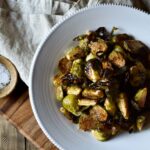 An overview of a bowl of Roasted Garlic Balsamic Brussel Sprouts on a napkin with a small bowl of salt nearby.