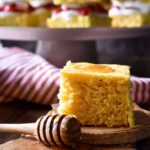 A piece of cornbread and honey with Cornbread Strawberry Shortcakes in the background.