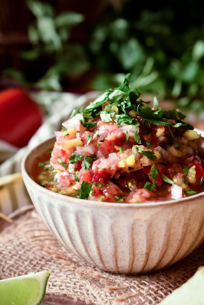 A bowl of Raul's Fresh Mango Salsa Fresca with cilantro, tomato and lime in the background.