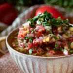 A bowl of Raul's Fresh Mango Salsa Fresca with chips, cilantro, tomato and lime in the background.