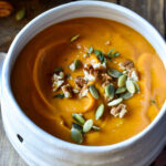 A close up of Sweet Potato Soup topped with pecans and pumpkin seeds in a mummy halloween bowl surrounded in fall leaves