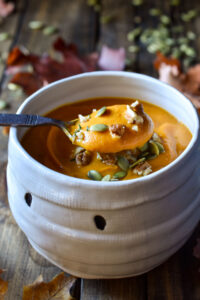 A close up of a spoon of Sweet Potato Soup topped with pecans and pumpkin seeds in a mummy halloween bowl surrounded in fall leaves